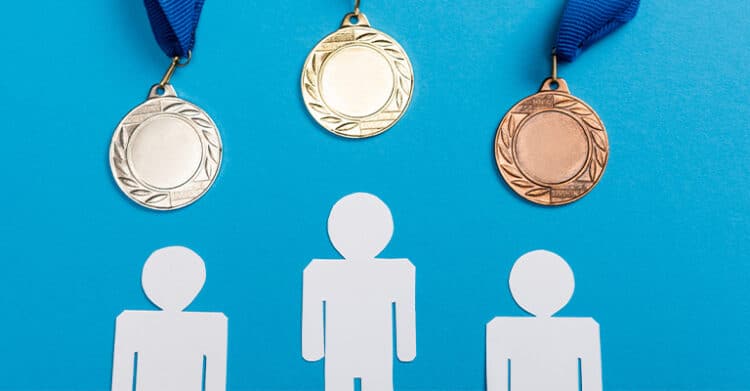 Three medals gold silver and bronze hang against a blue background above simple human figure icons. The image represents ranking achievement or comparison of performance such as winners first second and third place.