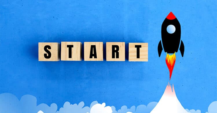Wooden letter blocks spell the word START on a blue background next to a cartoon style rocket blasting upward with flames. The image represents launching a new idea, beginning a project, or taking the first step toward growth or momentum.