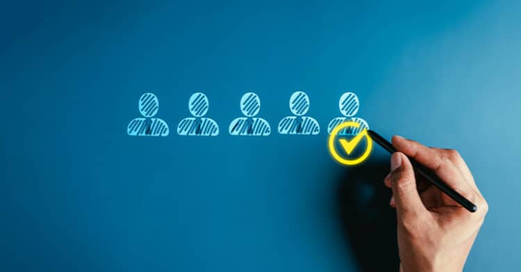 Audience research concept with a row of user icons on a blue background as a hand uses a pen to place a yellow check mark on one selected profile. The image highlights choosing the right audience segment during audience research.