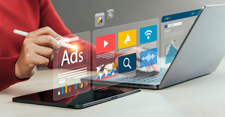 A person uses a tablet beside an open laptop while floating digital panels display marketing icons, charts, and the word "Ads." The scene supports digital strategy by showing online advertising, content distribution, and performance tracking across connected devices.