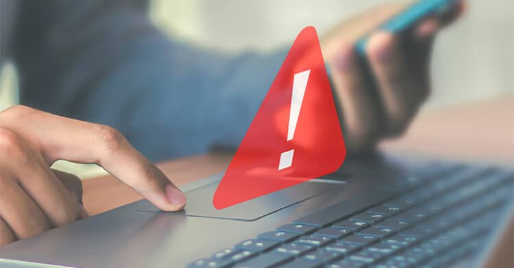 A person touches a laptop trackpad while holding a smartphone as a red warning icon with an exclamation mark appears above the keyboard. The image supports digital strategy by representing risk detection, urgent issues, and the need to respond quickly to problems across digital platforms.