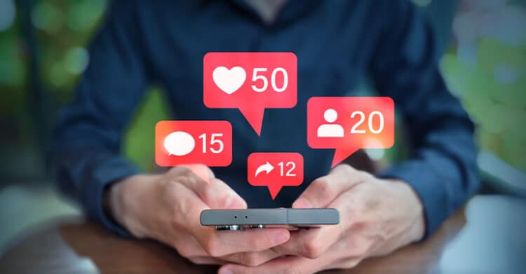 How to Build a Social Media Strategy That Works 7 Person holding a smartphone with floating notification icons showing "50" likes, "20" followers, "15" comments, and "12" shares above the screen. This visual represents a social media strategy by highlighting engagement metrics used to measure content performance.
