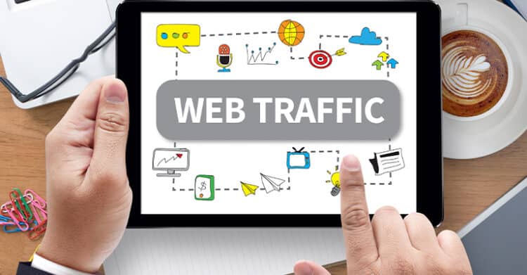 Person holding a tablet that reads WEB TRAFFIC with doodle style marketing icons around it like charts targets and paper airplanes. Coffee and desk supplies frame the scene to illustrate tips to Drive Traffic to Your Website.