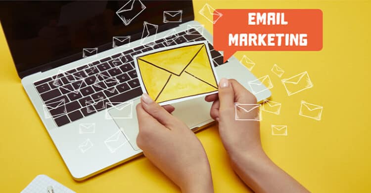Hands holding a yellow envelope in front of an open laptop on a bright yellow desk with illustrated envelope icons flying around. The image includes the text EMAIL MARKETING to represent sending campaigns and building an email list.