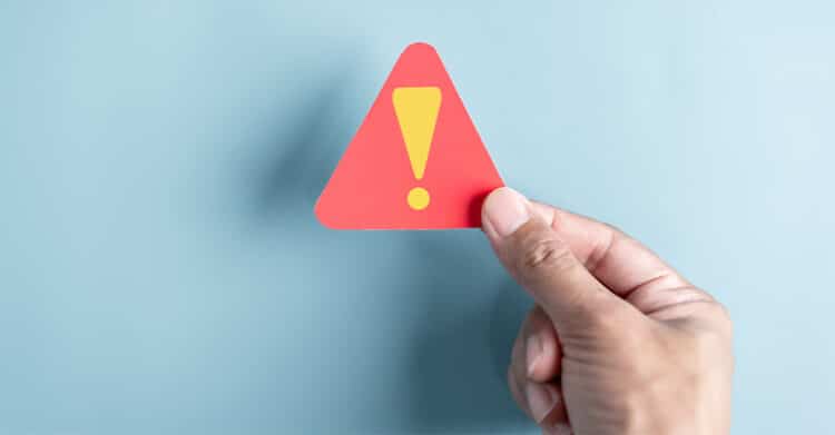 Hand holding a red triangular warning sign with a yellow exclamation mark against a light blue background. Simple visual that signals an important alert or something to watch out for.