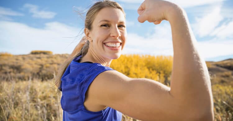 How to Write Blog Titles that Earn Clicks (A Complete Guide) 6 Smiling woman outdoors in a blue athletic top flexing her arm against a bright sky and dry grassy landscape. This visual can support a post about Blog Titles by suggesting strength, confidence, and powerful wording that helps a headline stand out.