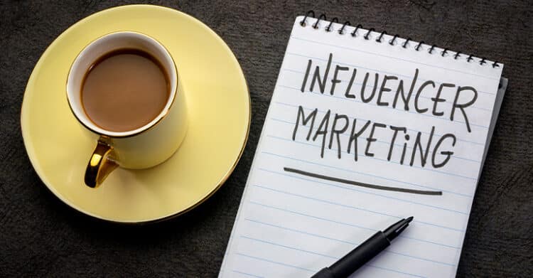 Coffee cup on a yellow saucer beside a spiral notebook with the words "INFLUENCER MARKETING" written in large letters and underlined. A black pen rests at the bottom of the notepad on a dark tabletop.