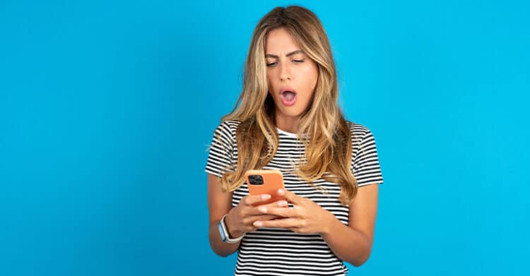 Woman in a black and white striped shirt looks shocked while reading her smartphone against a bright blue background. Her surprised expression suggests reacting to influencer marketing results like campaign performance or a sponsored post going viral.