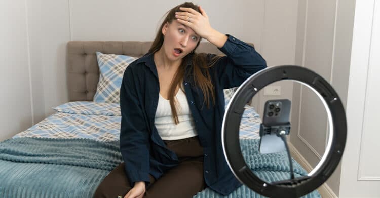 Woman sitting on a bed reacts with surprise while filming herself with a smartphone on a tripod inside a ring light. The behind the scenes setup shows an influencer marketing style video shoot in a bedroom.