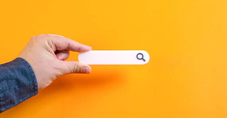A hand holds a blank search bar with a small magnifying glass icon against a bright yellow background. This simple graphic represents SEO strategy through the idea of search intent, keyword targeting, and online visibility.