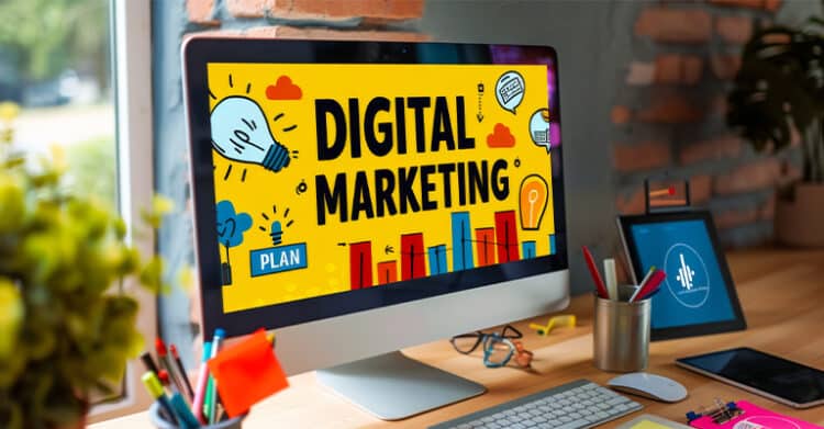 A desktop monitor on a workspace displays the words "DIGITAL MARKETING" in bold on a bright yellow screen with charts, light bulb icons, and a small label that says "PLAN." This scene supports SEO strategy by showing digital marketing as a broader effort that includes planning, content, and performance tracking.