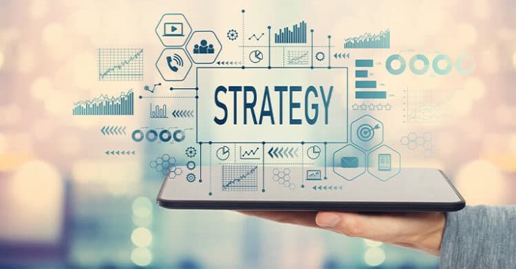 Hand holding a tablet with the word "STRATEGY" displayed in the center, surrounded by charts, graphs, icons, and performance dashboards on a blurred background. This concept graphic supports content about how to increase social media engagement by highlighting the importance of planning, analytics, and a clear content strategy.