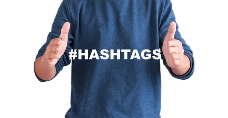 Person in a blue sweater holds their hands apart around the word "#HASHTAGS" on a plain light background. This graphic supports content about how to increase social media engagement by pointing to hashtags as a tool for expanding reach and helping posts get discovered.
