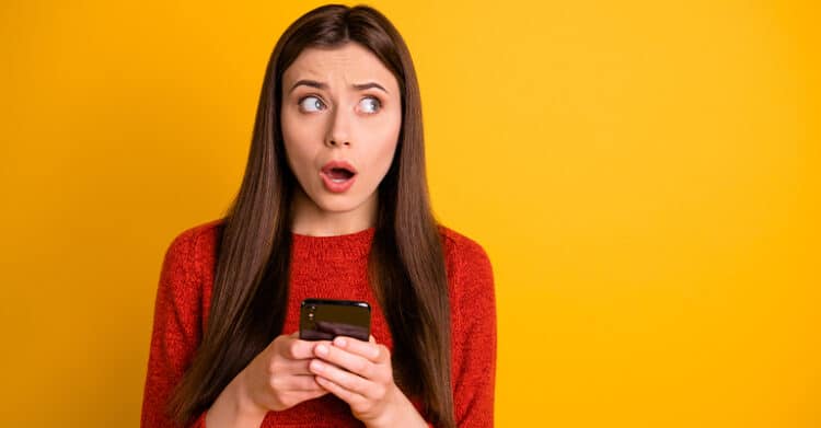 Woman in a red sweater stares sideways in surprise while holding a smartphone against a bright yellow background. This photo supports content about how to increase social media engagement by capturing the kind of strong reaction that eye catching posts can create.
