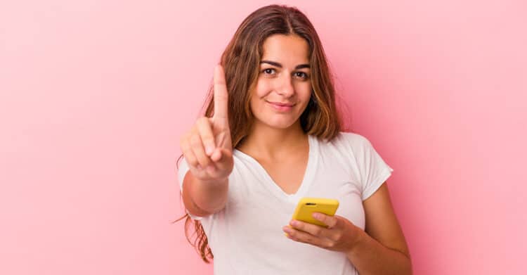Mobile-First Indexing: How to Prepare Your Website & Content 4 Woman holding a yellow smartphone and raising one finger while looking at the camera against a pink background, representing mobile-first indexing and prioritizing mobile users.