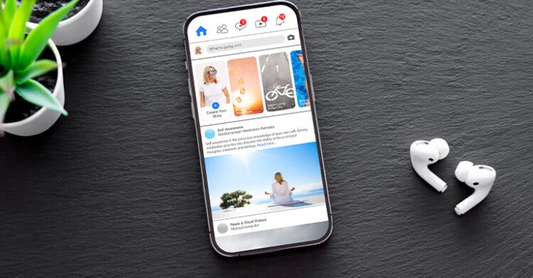 Smartphone displaying a social media marketing feed with posts and stories on screen, placed on a dark surface next to wireless earbuds and a green plant.