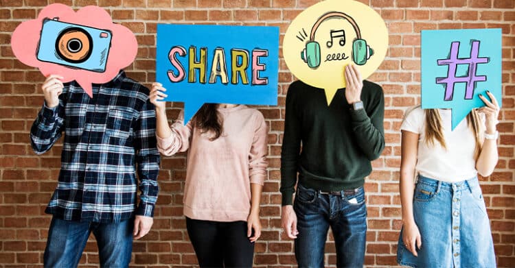 Four people standing against a brick wall holding colorful speech bubble signs with icons of a camera, the word "SHARE", headphones with a music note, and a hashtag symbol. Visual representation of social media rules emphasizing content sharing, hashtags, and multimedia communication online.