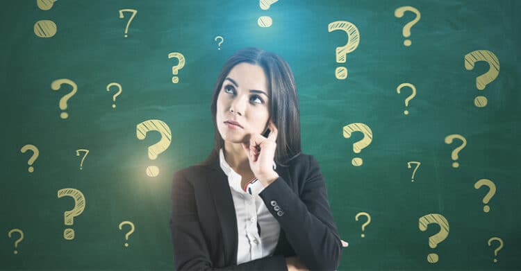 Woman in business attire stands in front of a green chalkboard filled with drawn question marks while looking up thoughtfully. Visual for social media rules emphasizing critical thinking and asking questions before posting or engaging online.