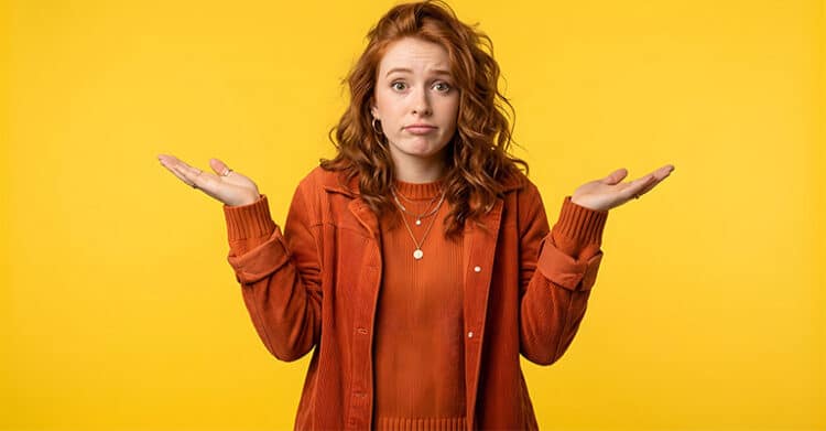 A red haired woman in an orange outfit shrugs with both hands raised against a bright yellow background. Her unsure expression suggests the confusion people may feel when figuring out how to create AI-Proof Content.