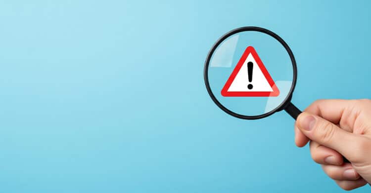 A hand holds a magnifying glass over a red warning symbol with an exclamation point on a blue background. The alert sign suggests checking for weak spots or risks before publishing AI-Proof Content.